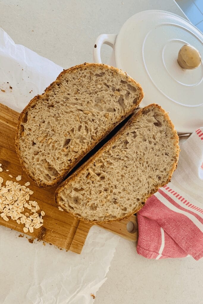 Loaf of multigrain sourdough bread that has been sliced open through the middle to show the inside of the bread. There is a cream enamel Dutch Oven and a red and white dish towel to the right of the cut loaf.