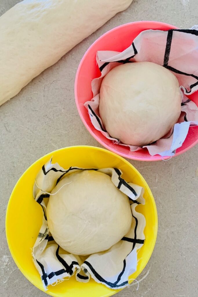 Yellow and pink plastic children's bowls lined with black and white fabric. Mini boules placed inside to proof.