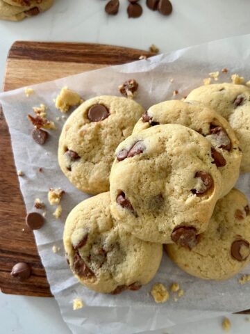 Pile of sourdough chocolate chip cookies sitting on a wooden serving board. There are chocolate chips sprinkled on the white benchtop.