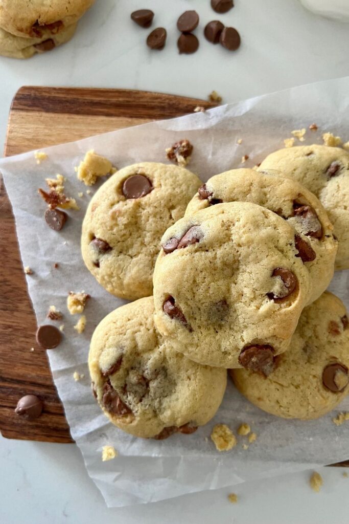 Pile of sourdough chocolate chip cookies sitting on a wooden serving board. There are chocolate chips sprinkled on the white benchtop.