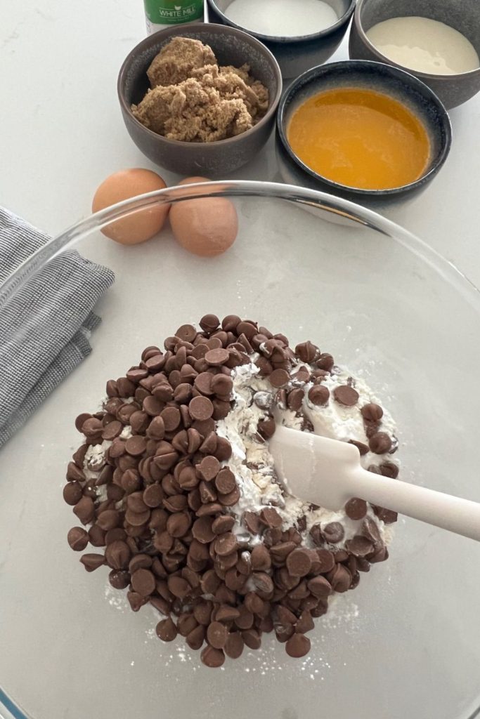 Glass bowl with dry ingredients in it, including chocolate chips. There are 2 eggs, brown sugar and melted butter in the background.