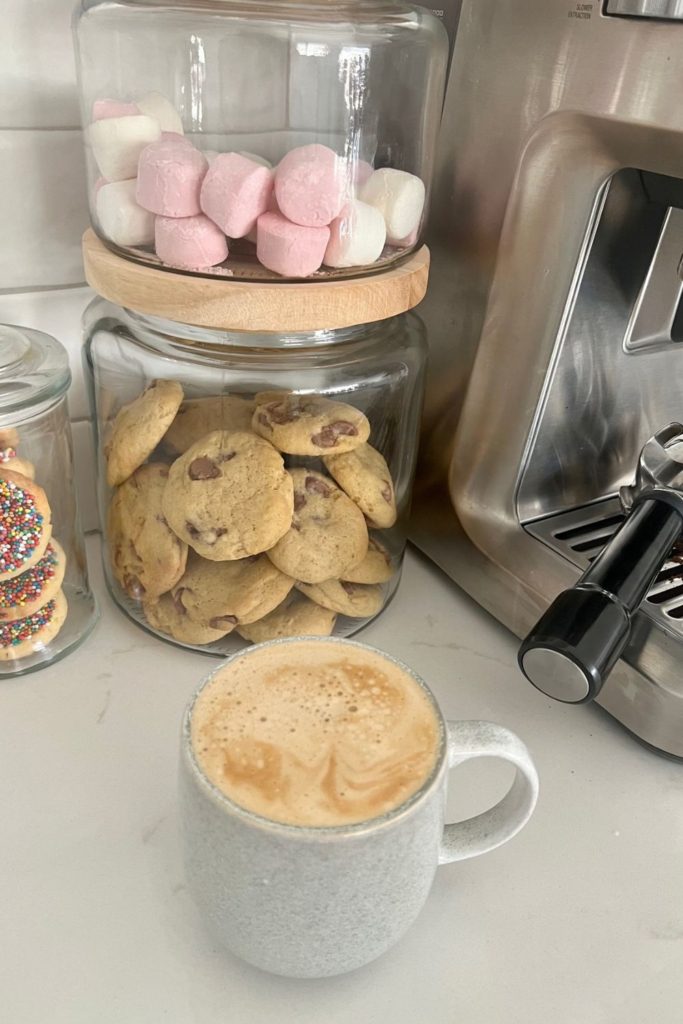 A cup of coffee sitting in front of a glass cookie jar with sourdough chocolate chip cookies and a jar of marshmallows.