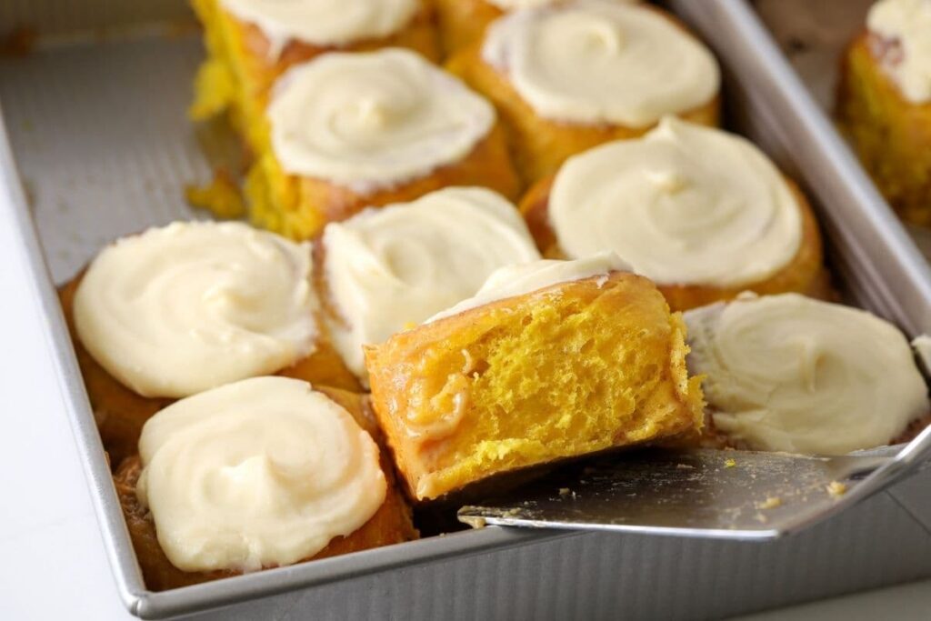 A tray of sourdough pumpkin cinnamon rolls that have been frosted in vanilla cream cheese frosting. One of the front rolls is being lifted up so you can see the fluffy, soft dough inside.