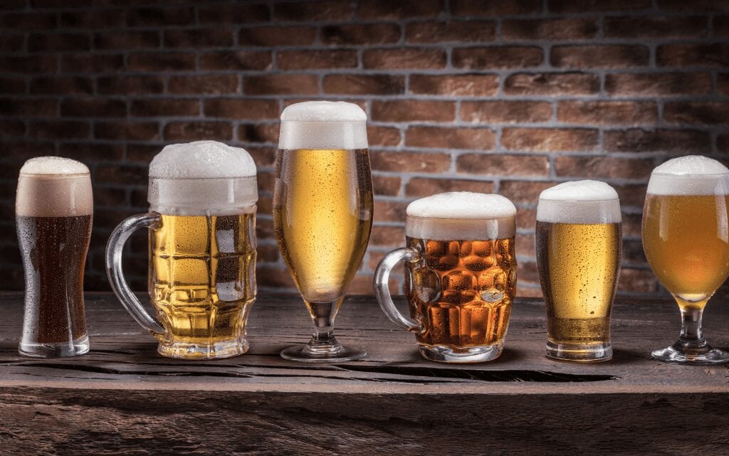 Six glasses of beer sitting in front of a brick wall.