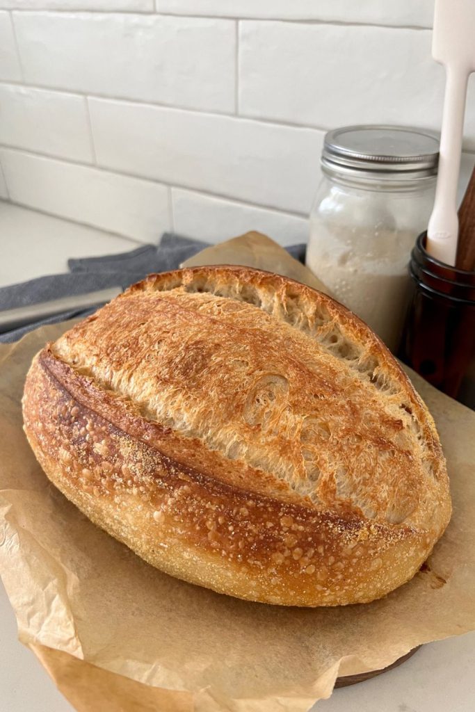 Loaf of sourdough bread that has been baked without Dutch Oven. It is sitting in front of a jar of sourdough starter.