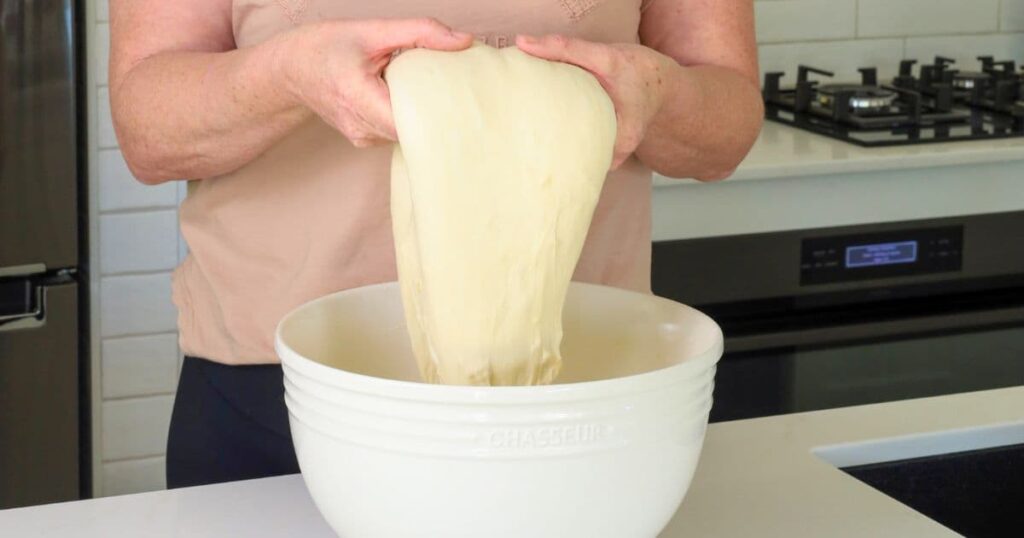 A photo showing a large cream colored bowl and someone performing coil folds on the dough out of the top of the bowl.