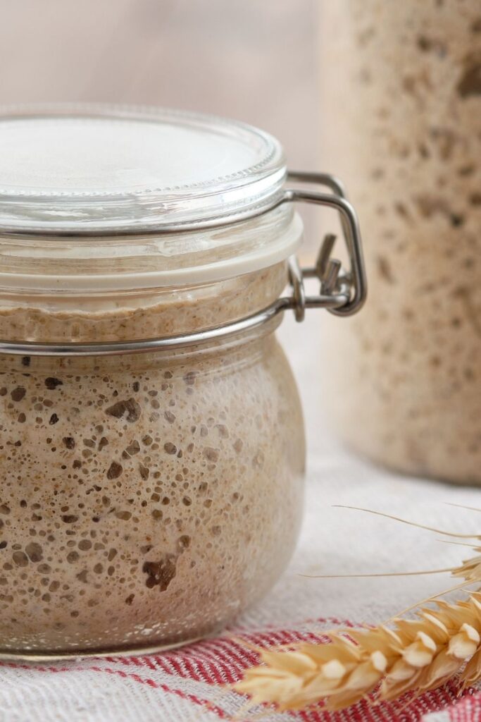 Rye sourdough starter shown from the side of the jar. There is a an ear of wheat and a red and white dish cloth in the photo too.