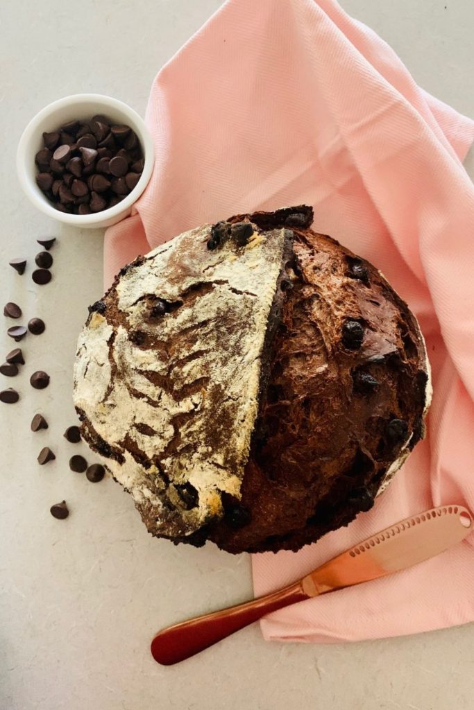 Loaf of chocolate sourdough discard bread sitting on a pale pink tea towel and surrounded by chocolate chips