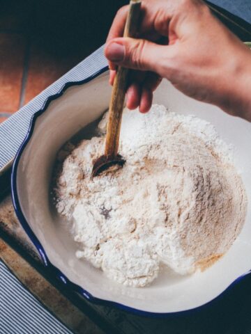 Wooden chopping board with a blue rimmed bowl sitting on top. The bowl is filled with flour and water and being stirred with a wooden spoon.