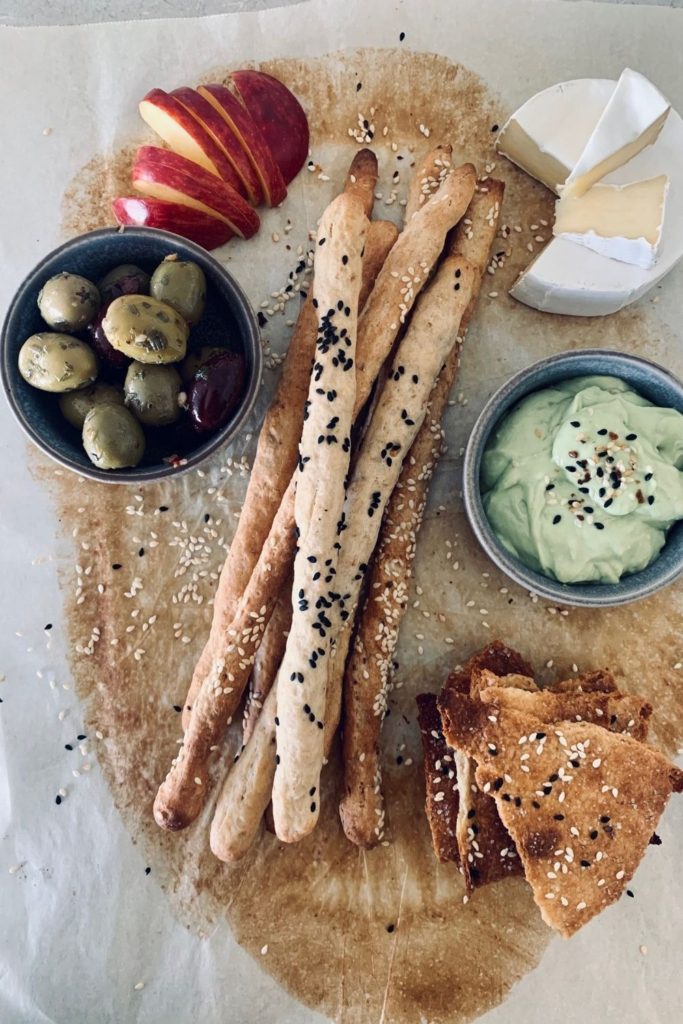 Sourdough grissini served in the middle of a charcuterie platter with green olives, red apple, sourdough lavosh crackers and avocado dip.