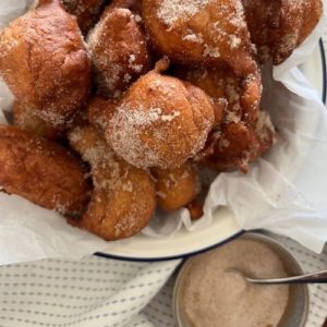 Cooked sourdough banana fritters dusted with cinnamon sugar. There is a small bowl of cinnamon sugar with a silver spoon sitting beside the fritters which are in a white and blue rimmed bowl.