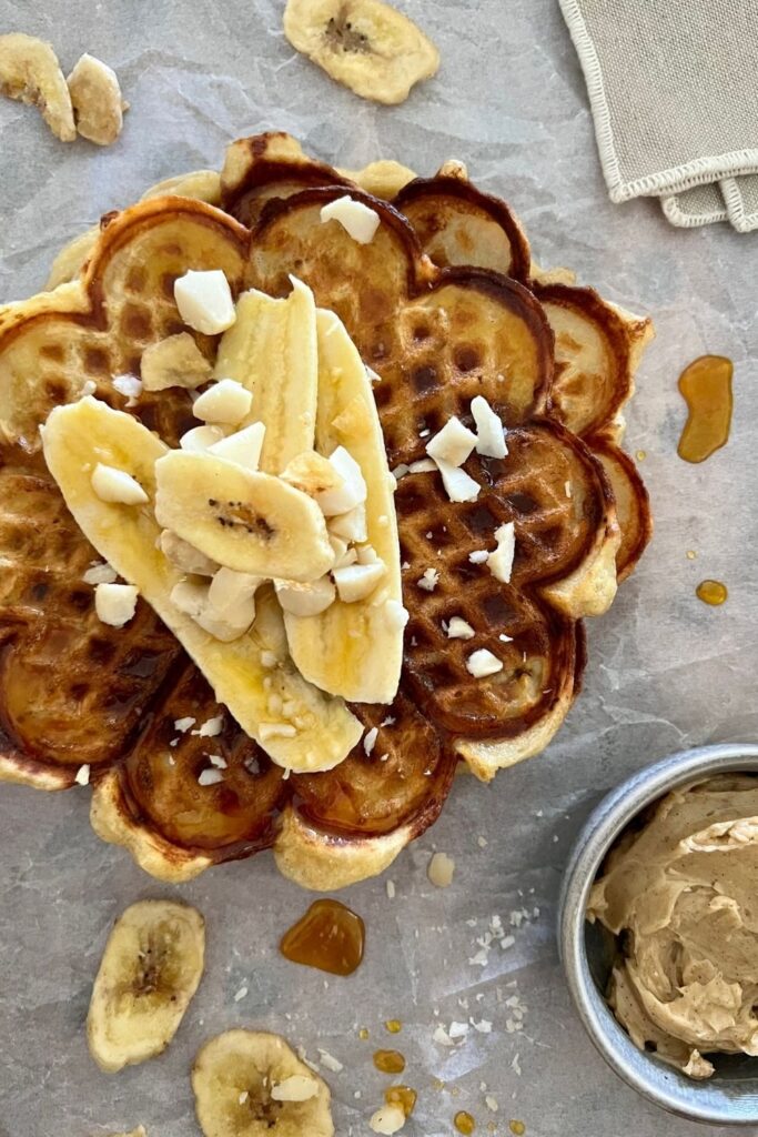 Sourdough banana waffles using sourdough banana fritter mixture. Waffles are served on paper, stacked and topped with sliced banana and crushed macadamia nuts.