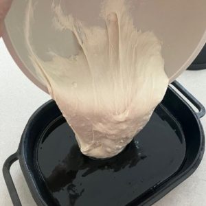 Pouring focaccia dough from a plastic bowl into a heavily oiled cast iron pan.