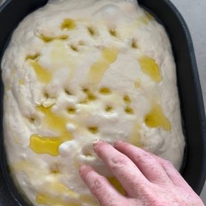 Sourdough focaccia dough drizzled in olive oil and being dimpled by fingertips.