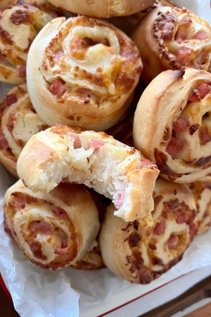 Sourdough pinwheels with ham and cheese piled up - there is one with a bite taken out of it to show the fluffy texture on the inside.