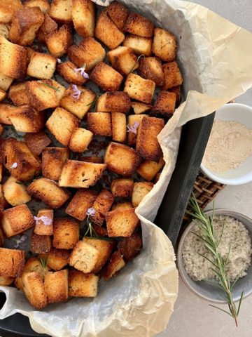 Crunchy homemade sourdough croutons in a cast iron dish lined with parchment paper. There is a bowl of french sea salt and sprig of rosemary to the right.