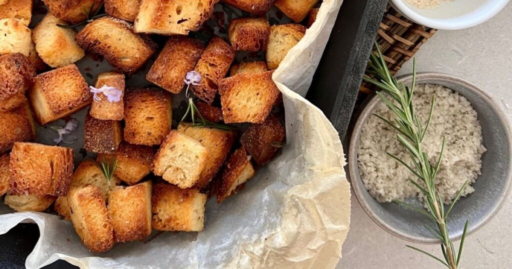 Crunchy sourdough croutons displayed in a cast iron bread pan lined with parchment paper. There is a dish of sea salt garnished with rosemary beside the pan of croutons.