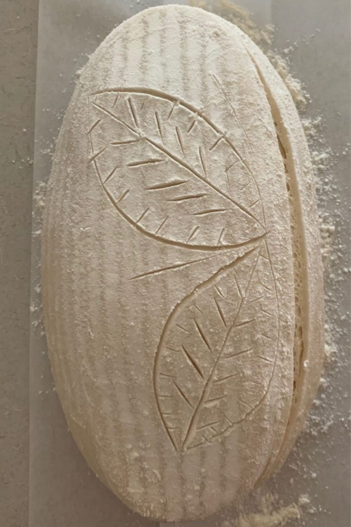Best tips for scoring sourdough bread - sourdough batard on a piece of parchment paper. It has one utility score on right hand side and two leaves scored on the left side.