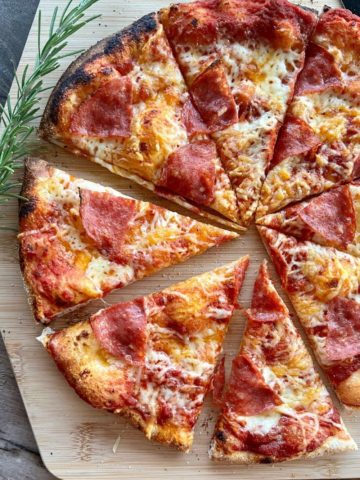 Sourdough pizza topped with pepperoni and cheese and sitting on a wooden pizza peel. There is a sprig of roseary on the left side of the pizza.