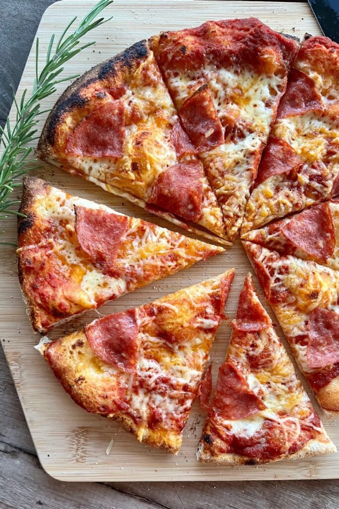 Sourdough pizza topped with pepperoni and cheese and sitting on a wooden pizza peel. There is a sprig of roseary on the left side of the pizza.