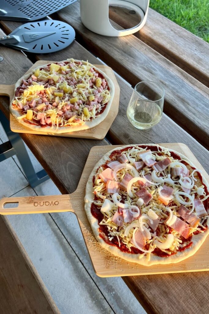 2 uncooked sourdough pizzas sitting on pizza peels. There is a glass of wine in the middle of them.