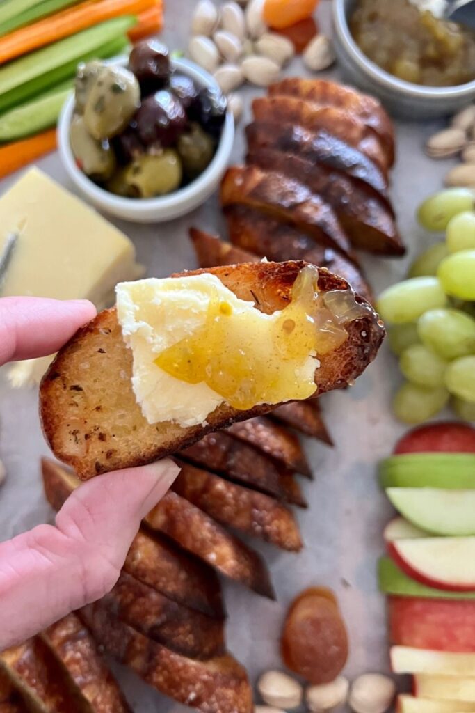 Close up of sourdough crostini topped with vintage cheddar and relish. You can see a charcuterie board in the background with olives, chese and grapes and apple.