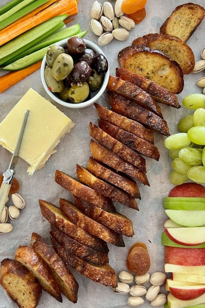 Sourdough Crostini are laid in a line through the middle of a charcuterie board featuring cheese, olives, relish, fresh fruit and vegetables and dried fruits and nuts.