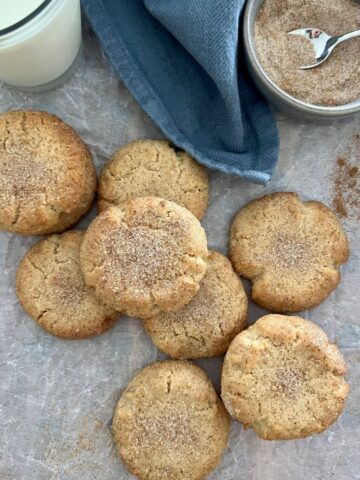 Perfect sourdough snickerdoodle cookies on a white background. There is a glass of milk, blue cloth and bowl of cinnamon sugar in the top half of the photo.