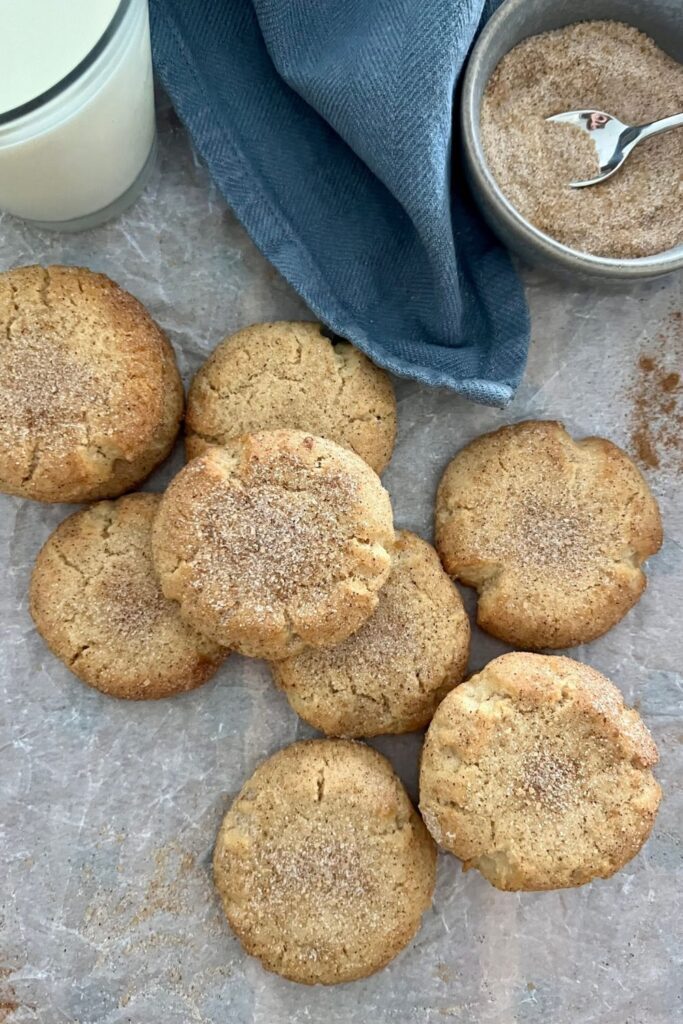 Perfect sourdough snickerdoodle cookies on a white background. There is a glass of milk, blue cloth and bowl of cinnamon sugar in the top half of the photo.