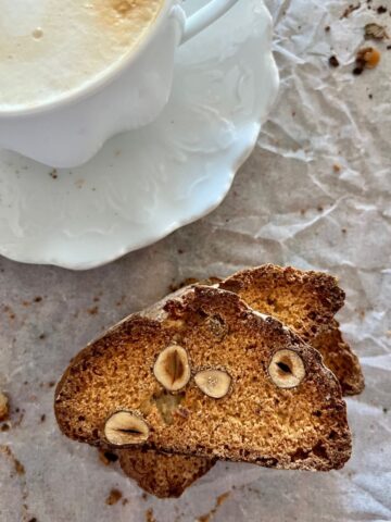 Sourdough biscotti stacked on a white background. There is a coffee cup displayed in the top left of the photo.