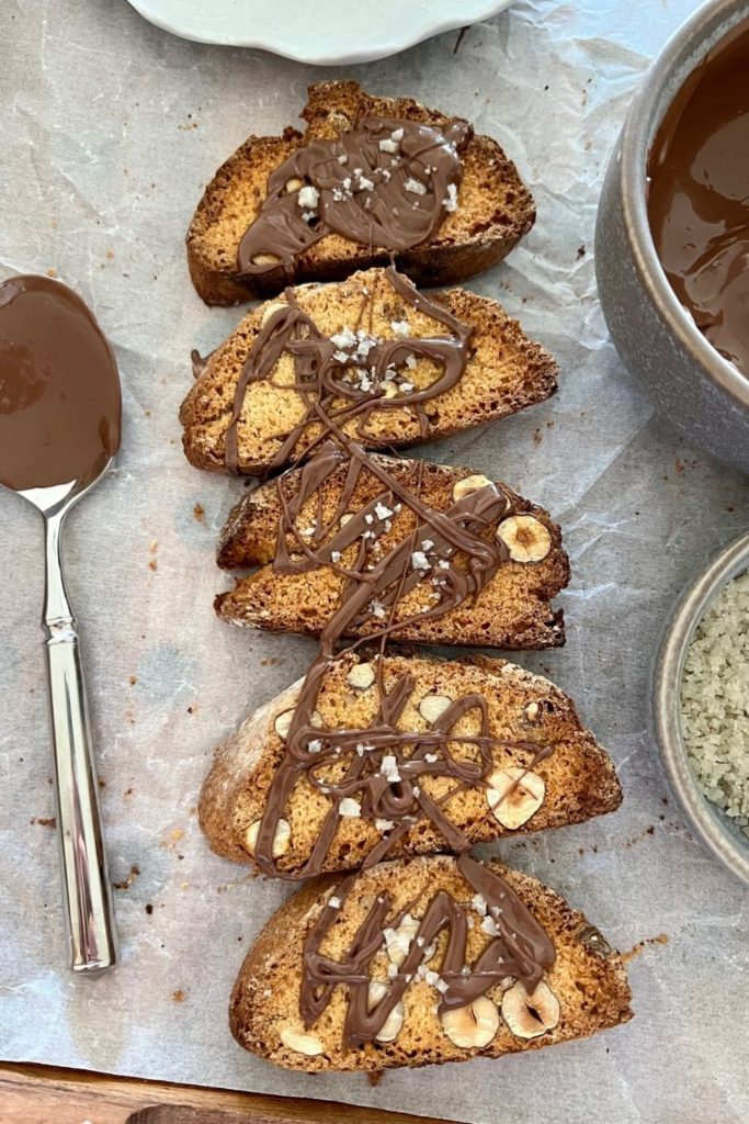 Sourdough biscotti drizzled with melted chocolate and sprinkled with sea salt. There is a spoon on the left hand side of the biscotti and you can see the melted chocolate and sea salt flakes in a bowl on the right.