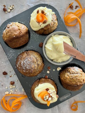 Sourdough carrot cake muffins in a six cup muffin tray. Two of them are topped with cream cheese frosting and carrot garnish.