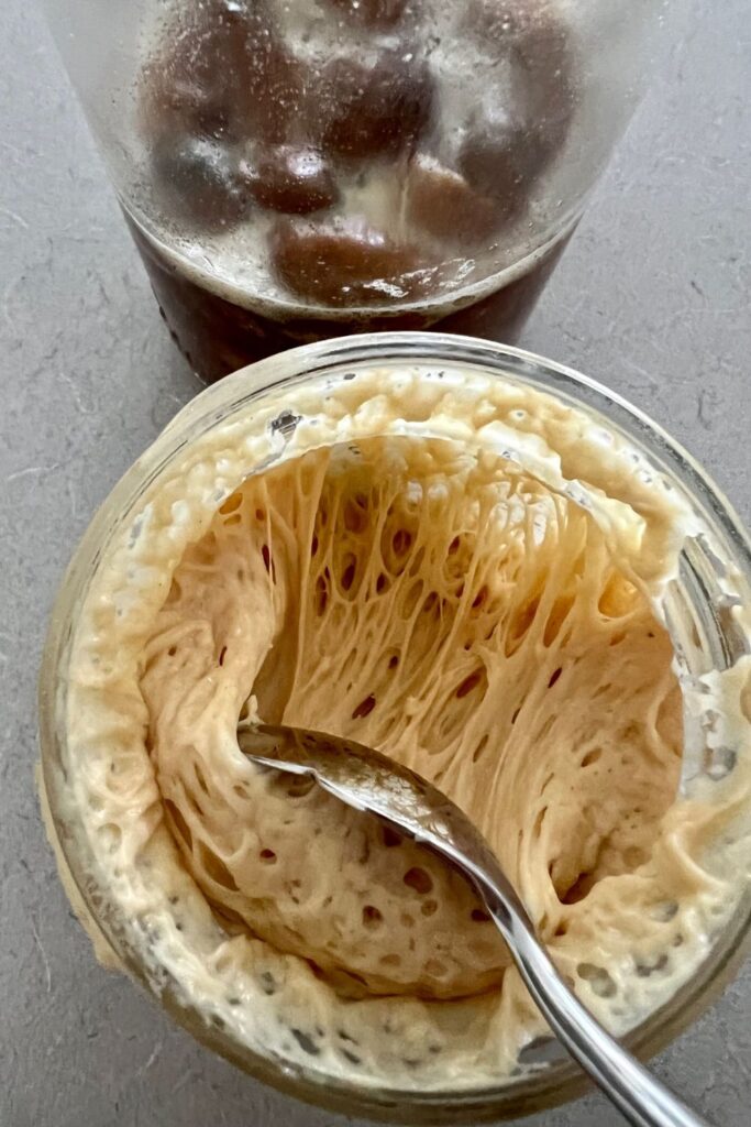 Jar of yeast water made with dried dates sitting behind a jar of sourdough starter made with the yeast water. There is a spoon in the sourdough starter to show the stretchiness of the starter.