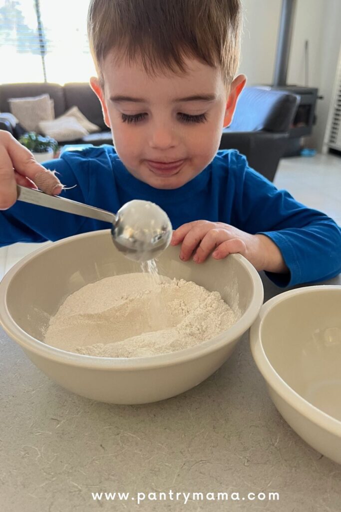 This is how busy moms make sourdough bread - involve the kids in the process. A 3 year old child in a blue shirt is measuring flour into a bowl.