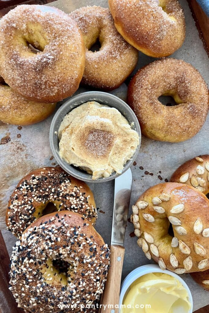 Tray of sourdough pumpkin bagels. The ones at the top have been dipped in cinnamon sugar. The ones on bottom left have been dipped in Everything Bagel Seasoning and the ones on the bottom right are topped with dried pumpkin seeds.