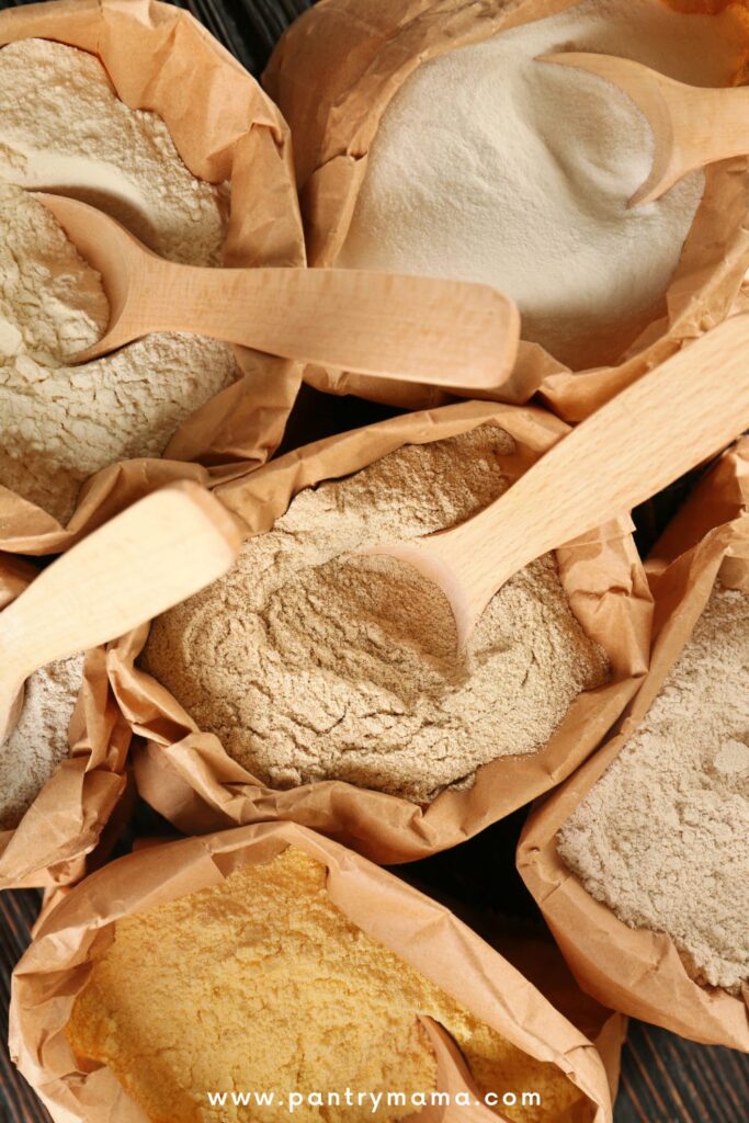 Brown paper bags containing different types of flours for sourdough starters. There is a wooden spoon in each bag of flour.