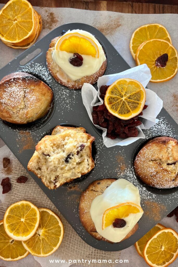 Tray of orange cranberry sourdough muffins with one of the muffins cut in half to show the tender crumb. 2 of the muffins are topped with vanilla cream cheese frosting.