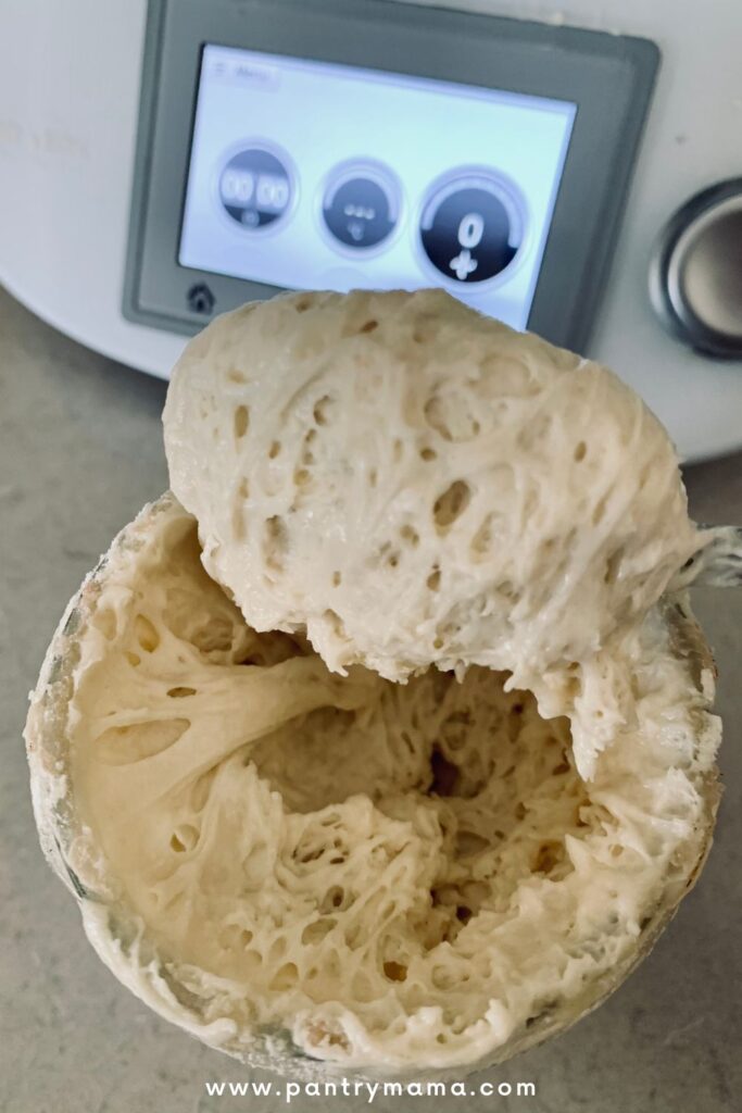 Thick sourdough starter being spooned out of the jar. There is a Thermomix in the background.