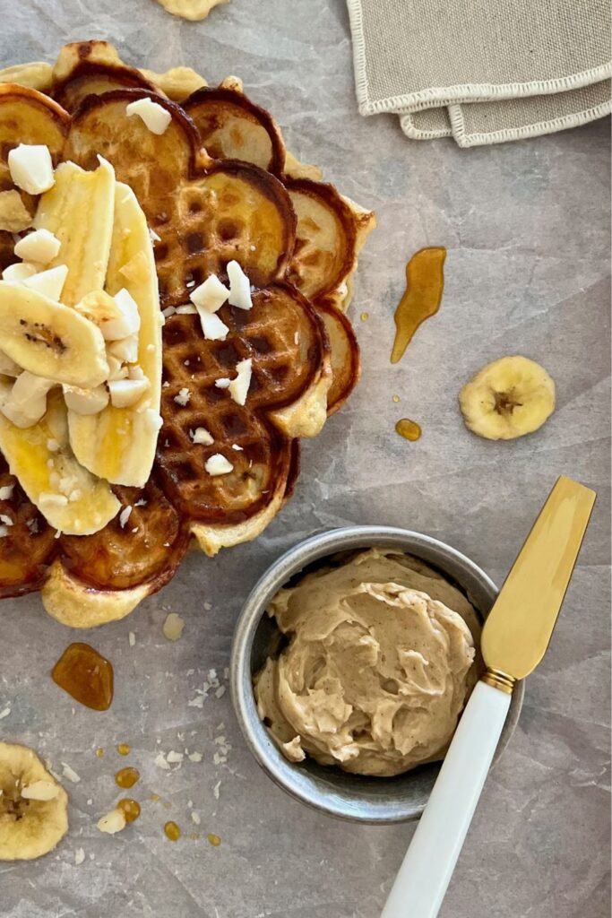 A small grey dish of whipped honey cinnamon butter with a gold knife sittlng on the edge. There is a stack of sourdough banana waffles sitting next to the dish.