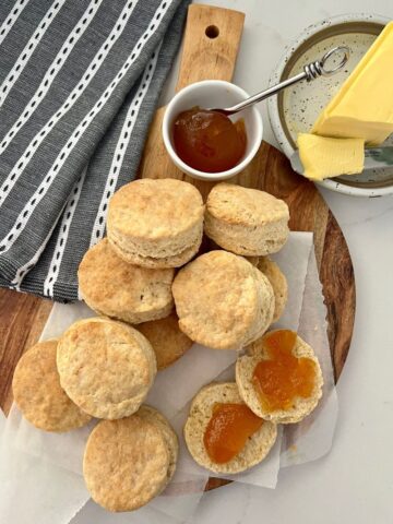 Buttery and flaky sourdough biscuits displayed on a wooden paddle smothered in apricot jam and butter.