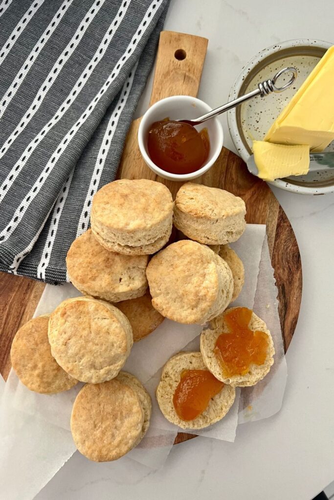 Buttery and flaky sourdough biscuits displayed on a wooden paddle smothered in apricot jam and butter.