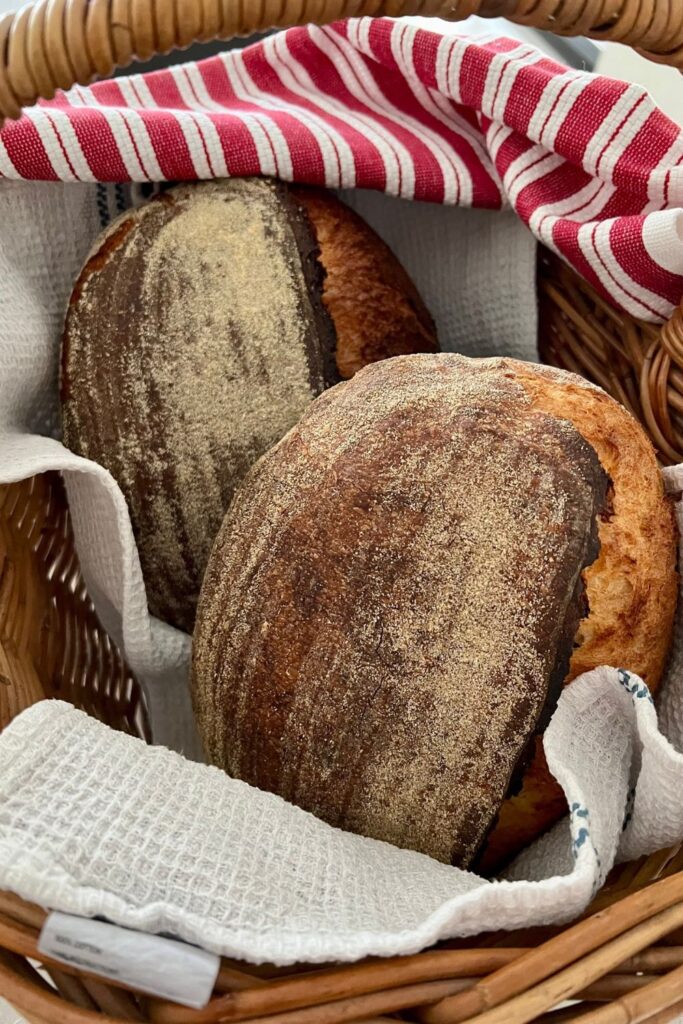 2 loaves of sourdough bread in a cane basket. They are partially wrapped in a red and white dish towel.