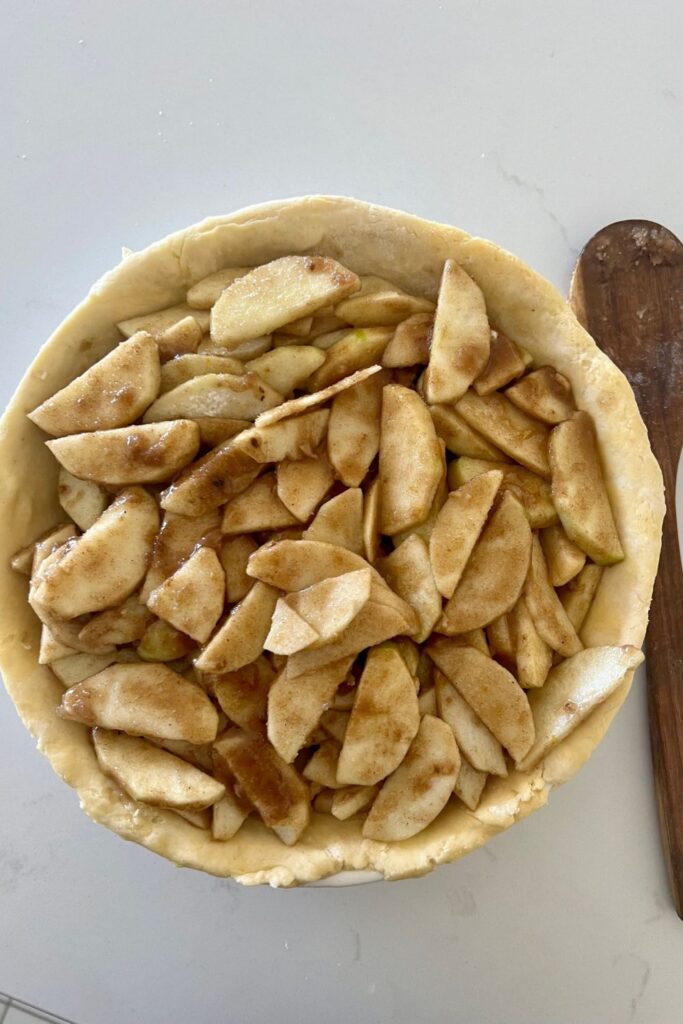 Pie dish lined with sourdough pie crust and filled with apple mixture. There is a wooden spoon sitting to the right of the sourdough apple pie. The pie has no top crust.