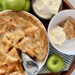 Sourdough apple pie displayed on a round wooden tray. A slice has been cut from the apple pie and there is a silver pie slice sitting in the dish. The dish is surrounded by 5 green granny smith apples, a bowl of whipped cream and a dish with a serve of apple pie and whipped cream.