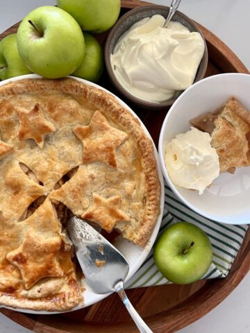 Sourdough apple pie displayed on a round wooden tray. A slice has been cut from the apple pie and there is a silver pie slice sitting in the dish. The dish is surrounded by 5 green granny smith apples, a bowl of whipped cream and a dish with a serve of apple pie and whipped cream.