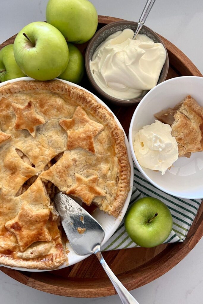 Sourdough apple pie displayed on a round wooden tray. A slice has been cut from the apple pie and there is a silver pie slice sitting in the dish. The dish is surrounded by 5 green granny smith apples, a bowl of whipped cream and a dish with a serve of apple pie and whipped cream.