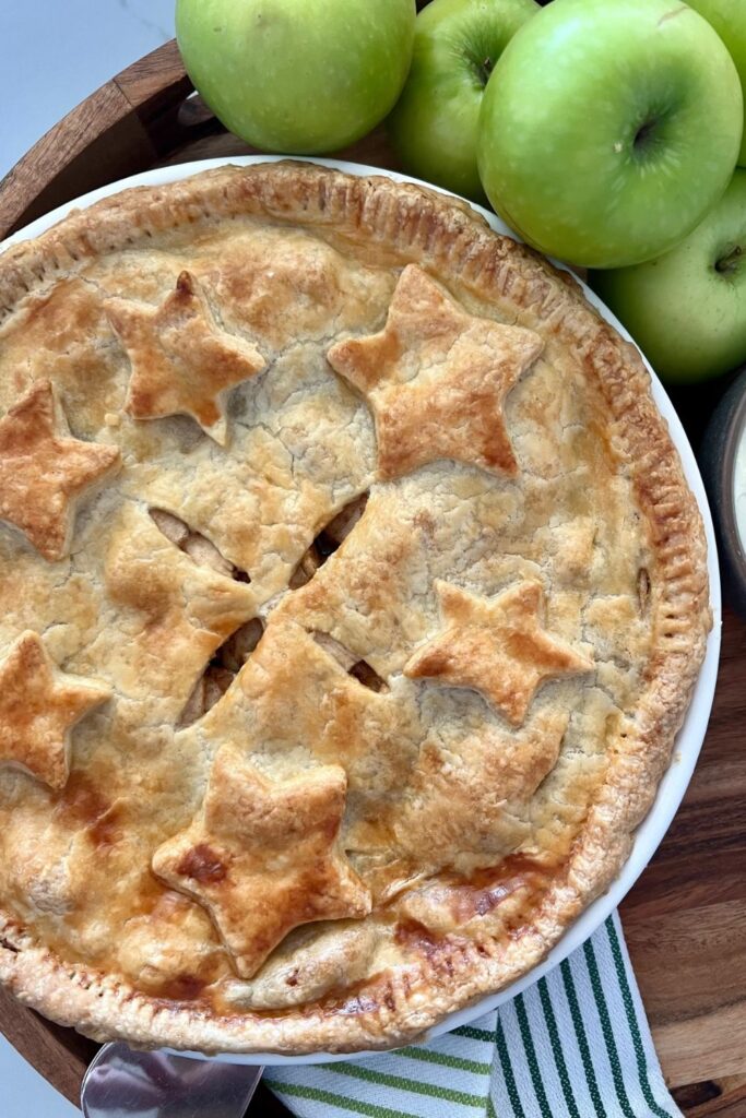 A whole, baked sourdough apple pie sitting in a white pie dish. The pie is surrounded by green granny smith apples and a green and white striped dish towel.