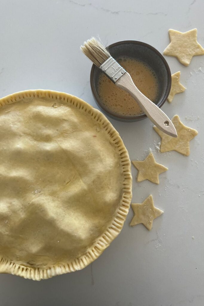 An unbaked sourdough apple pie that has been egg washed. There is a pastry brush and dish of egg wash sitting to the right of the pie as well as star shapes that have been cut out of excess sourdough shortcrust pastry with a cookie cutter.