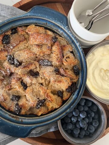 Sourdough bread pudding with raisins baked in a blue casserole dish. It is on a wooden tray with a dish of whipped cream and a bowl of blueberries.