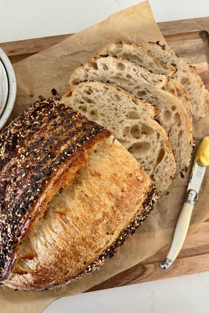 Loaf of Everything Bagel Seasoning Sourdough that has had 3 slices cut from it. There is an ivory handled knife with butter on it sitting to the right of the bread.